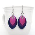 Handmade chainmail earrings featuring layered anodized aluminum scales in bisexual pride colors – hot pink, purple, and blue – hanging from a white ceramic mug.