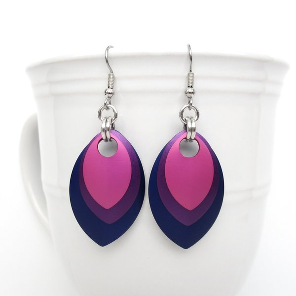 Handmade chainmail earrings featuring layered anodized aluminum scales in bisexual pride colors – hot pink, purple, and blue – hanging from a white ceramic mug.